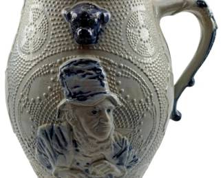 93 - Antique Salt Glazed Stoneware Pitcher (Description): Antique salt glazed stoneware pitcher featuring a relief portrait of a gentleman in period attire, accented with cobalt blue highlights. The body displays textured beaded patterns and decorative medallions, with a cobalt blue dog head ornament below the spout. The graceful handle features additional blue detailing. Measures 11" x 8" x 5".