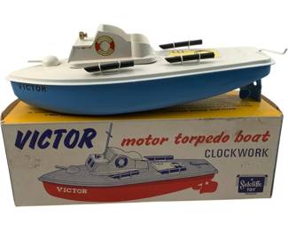 1185 - Sutcliffe Victor Motor Torpedo Boat w/ Box (Description): Sutcliffe Victor Motor Torpedo Boat, a clockwork-powered tinplate toy in blue and white livery. The boat features detailed cabin structure, twin torpedoes, life ring detail, and rear-mounted propeller mechanism. Comes with its original yellow and white illustrated box. A classic British-made toy boat from the Sutcliffe company.