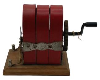 864 - Antique Hand Crank Generator 4.25"x4"x10" (Description): Antique hand crank generator featuring a red painted metal housing with three cylindrical coils secured by a decorative metal band. The unit is mounted on a wooden base with brass binding posts and coiled wire connections. A black bakelite hand crank operates the mechanism via a brass gear wheel. Measures 4 1/4" x 4" x 10".