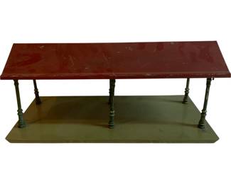 1163 - Metal Train Depot 8"x14"x9" (Description): Metal train depot featuring a red-painted roof supported by six turned green metal posts on a green rectangular base. The piece shows desirable age-related patina and paint wear consistent with vintage use. Measures 8 inches by 14 inches by 9 inches.
