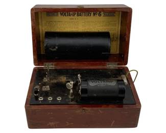 1094 - Voltamp Battery #6 Majestic in Wood Box (Description): Voltamp Battery No. 6 "Majestic" housed in its original mahogany-stained wooden case. The lid interior features printed directions for operation and includes the serial number. The device includes the black cylindrical coil marked "Voltamp" and ml contact mechanisms mounted on a black base plate. The case shows appropriate age wear consistent with vintage electrical equipment. Measures 5 1/2" x 9" x 5.5".