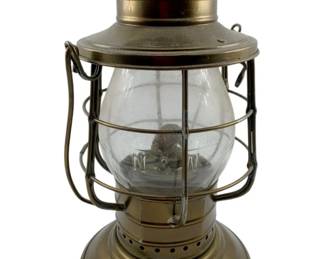 91 - Universal Brass Railroad Lantern (Description): Universal Metal Spinning & Stamping Co brass railroad lantern featuring a clear glass globe protected by a wire cage guard. The lantern has a ventilated dome top, brass construction throughout, and a sturdy base with perforated detailing. The globe appears to have embossed lettering. Measures 12" x 6.5".