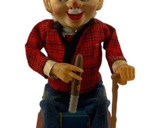 924 - Smoking Charlie Battery Operated Toy (Description): Smoking Charlie battery operated toy featuring a seated elderly gentleman figure with a vinyl head, blonde hair, and bright red beret. He wears a red and black plaid jacket with black vest, red bow tie, and blue corduroy pants with black shoes. The figure holds a cigar in one hand and a cane in the other, seated on a blue tin box atop a red lithographed tin base. A classic Japanese novelty toy from the mid-century era.