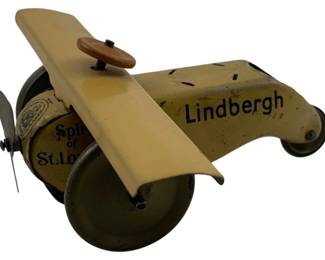 923 - Lindbergh Spirit of St. Louis Tin Litho Toy (Description): Tin lithographed metal toy airplane commemorating Charles Lindbergh's historic Spirit of St. Louis aircraft. This vintage toy features a cream and tan color scheme with black lettering reading "Lindbergh" on the fuselage and "Spirit of St. Louis" the engine cowling. The airplane includes a brown propeller disc, metal wings, and wheeled landing gear. The piece shows its age with gentle patina consistent with a toy from the