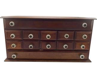 128 - Farmhouse 12 Drawer Apothecary Cabinet (Description): Rustic farmhouse style apothecary cabinet featuring twelve small drawers with porcelain knob pulls. The cabinet is crafted from dark stained wood with a warm mahogany finish. The piece includes two long horizontal drawers at top and bottom with ten smaller square drawers arranged in the center section. The cabinet sits on a molded base and features a beveled top edge. Measures 9.5" x 19" x 10".