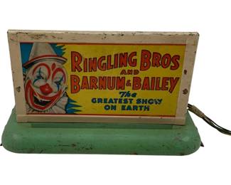 1187 - American Flyer Ringling Bros. Sign (Description): American Flyer Ringling Bros. and Barnum & Bailey circus sign toy featuring colorful lithographed tin construction. The sign displays a cheerful clown face alongside the iconic "The Greatest Show on Earth" slogan in red, yellow, and blue graphics. The sign panel sits atop a green painted metal base with an attached electrical cord, suggesting this functioned as an illuminated display piece. Shows vintage wear consistent with age