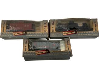 1129 - Gilbert HO Scale Trains in Boxes 3pc Lot (Description): A three-piece lot of Gilbert HO scale model trains, each in its original box. The collection includes a Reading caboose in maroon red, a black steam locomotive, and a gray hopper car. All boxes feature the classic Gilbert HO 'Top Line in HO' packaging with the 'Super Detailed - Ready to Go!' tagline. The trains and boxes show vintage wear consistent with age.