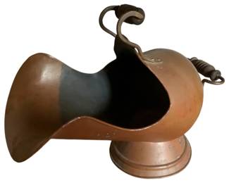 155 - Vintage Copper Coal Scuttle 16"x20"x13" (Description): Vintage copper coal scuttle featuring a classic helmet-style design with a wide flared opening for easy filling. The piece has a curved carrying handle on top and a secondary fixed handle on the back, both with wrapped grip details. Resting on a stepped circular base, this coal scuttle displays a warm copper patina consistent with age and use. Measures 16 inches by 20 inches by 13 inches.