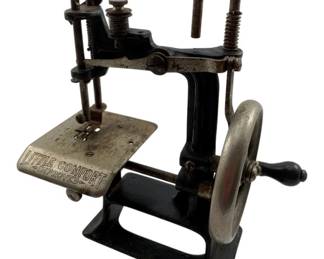 17 - Little Comfort Improved Mini Sewing Machine (Description): Little Comfort "Improved" mini sewing machine featuring a black cast iron body with nickel-plated accents. This antique hand-crank sewing machine has the "Little Comfort Improved" name embossed on the needle plate. The machine features a wooden handle, adjustable tension mechanism, and table clamp for securing to a work surface. Shows aged patina consistent with its vintage. Measures 7" x 6.5" x 3".