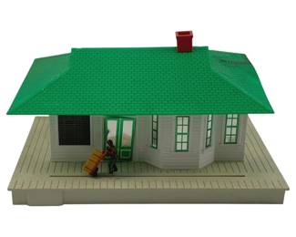 860 - American Flyer Lighted Station Toy (Description): American Flyer lighted station toy featuring a white clapboard building with a green shingled roof and red chimney. The station includes a wraparound covered platform with a figure pushing a hand truck loaded with cargo. Windows feature green trim and the structure includes detailed architectural elements. Measures 6" x 10" x 14". Used as is electric/electronic/mechanical item. No guarantee all components, untested.