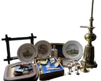 185 - Group Lot of Brass, Plates & Frames (Description): A mixed group lot featuring brass items, decorative plates, and frames. The lot includes a tall brass candlestick or lamp base with turned details, several small brass or silver-plated goblets, decorative collector's plates with scenic designs, a dark wooden trundle-style frame, lacquered serving trays, a child's tea set in original box, flatware, and various decorative accessories. A diverse collection suitable
