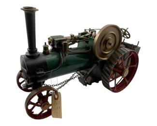 20 - Bassett-Lowke Live Steam Tractor Model (Description): Bassett-Lowke live steam tractor model featuring a green painted boiler with black firebox, brass flywheel, and red spoke wheels with metal cleats on the rear drive wheels. The model includes detailed brass fittings, a tall black smokestack, and operational steering mechanism with chain linkage. Measures 8 1/2" x 15" x 16".