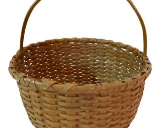 79 - Medium Primitive Basket w/Handle 14"x12.5" (Description): Medium primitive basket with handle, featuring traditional splint weave construction. The basket displays a natural light brown patina consistent with age and use. A sturdy bent wood swing handle is attached at the rim for carrying. Measures 14 inches by 12.5 inches.