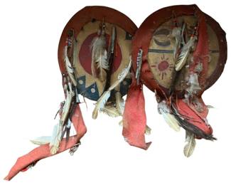 160 - Pr Vtg Native American Medicine Shields (Description): Group of two vintage Native American medicine shields featuring painted rawhide with traditional symbolic designs including sun and moon motifs. Each shield is decorated with feathers, beadwork, and red leather or hide embellishments. The painted surfaces display red, blue, and tan coloring with geometric patterns. Measures 32 inches by 18 inches.