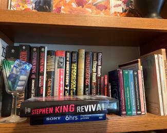 Stephen King: The Bachman Books, Stephen King: Drawing of the Three, Stephen King: The Dark Tower, Stephen King: The Gunslinger, Shark, Jagger (Christopher Andersen), New Dimensions 7, Winning Golf: The Professional Way, (two green/gold volumes – title obscured), Urban Legends (Jan Harold Brunvand), More Chicago Haunts, Chicago Haunts, The Rolling Stones (small book), Stephen King: Revival, New Webster’s Dictionary, New Webster’s Word Finder, New Webster’s Dictionary, New Webster’s Everyday Dictionary, New Webster’s Crossword Dictionary, New Webster’s Rhyming Dictionary, TV Guide, Endurance (Alfred Lansing), Alfred Lansing (partially obscured title), The Essential Grandma Cookbook (partially visible), The American Medical Association Family Medical Guide (partially visible), Sony Premium Grade T-120 VHS tape, glass with golf tees.