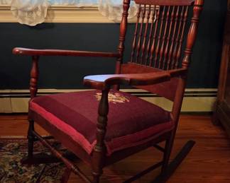 Red Mahogany Rocking Chair with needlepoint seat – 11 turned spindles, double serpentine and voluted crest with scrolled gold stenciled scroll, flat shaped arms with modified knuckles, turned legs single box stretcher circa 1890