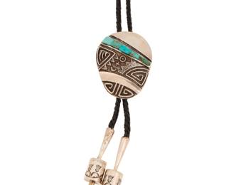 2244 A Harold Lovato Kewa/Santo Domingo Pueblo cast silver and turquoise bolo tie Harold Lovato (1954 - 1997, Kewa) Late 20th century Marked: HL / [cornstalk] A cast sterling silver oval bolo with geometric motifs in silver overlay and a line of turquoise inlay, with similar motifs to reverse, and all on a braided black leather cord with cylindrical barrel bead stamped silver tips with 'blossom' ends Bolo: 2.375" H x 1.25" W"; cord: 44" L 84.5 grams gross Estimate: $400 - $600