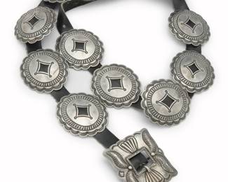 2096 A Fred Thompson Navajo sterling silver First Phase-style concho belt Fred Thompson (1925-2002, Diné) Mid-20th century With artist's tilde mark to buckle verso Comprising a chunky concho belt with a rectangular repoussé buckle and eleven oval conchos with stampwork details and scalloped edges, all mounted to a narrow black leather belt strap Buckle: 2.25" H x 2.5" W; Each oval concho: 2.25" H x 2.625" W; Belt: 41.5" L x 0.75" H 345.1 grams gross Estimate: $1,000 - $1,500