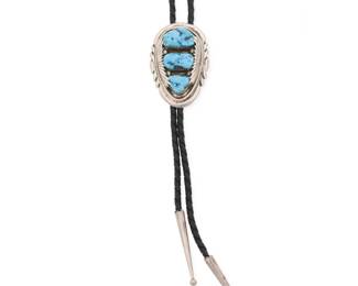 2245 A Robert & Bernice Leekya Zuni sterling silver and turquoise bolo tie Robert and Bernice Leekya (b. 1934 and b. 1930s, Zuni Pueblo) Late 20th/21st century Stamped: RLB An oval sterling silver bolo slide with graduated set turquoise and incised edging detail, suspended on a black braided leather strap with tapered silver tips Bolo slide: 2.25" H x 1.5" W; cord: 37" L 39.7 grams gross Estimate: $200 - $400