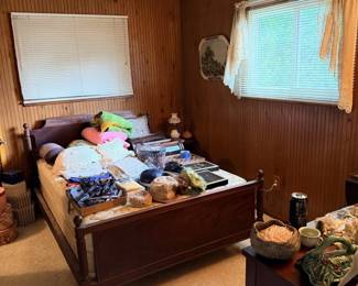 Bedroom overview with wood bed frame, bedding, baskets, folded clothing and small estate items throughout the room.