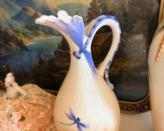 Tall white pitcher or vase with blue flower, displayed with shell/pottery and landscape art.