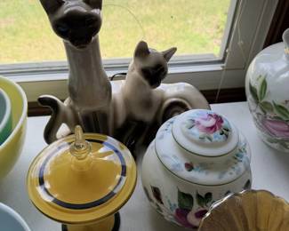 Siamese cat figures, floral lidded jars and yellow/white dishware near window.