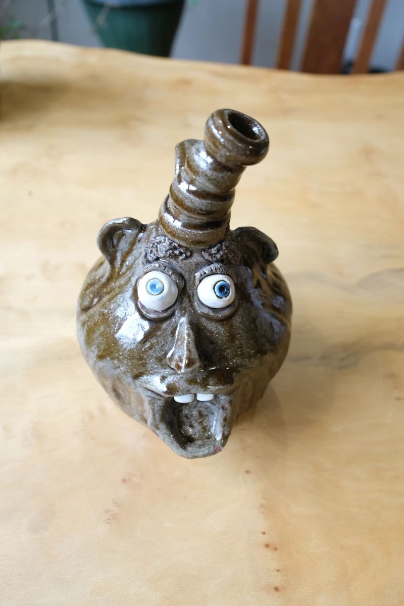 Up for sale is a highly collectible, authentic Southern folk art stoneware ceramic face jug handcrafted and signed by regional potter P. Hosey (Phillip Hosey) out of Carrollton, Georgia.This whimsical alkaline-glazed clay piece features an exceptionally unique, stylized smoking pipe or stack spout protruding from the top of the head, acting as a secondary vessel handle or opening. The figure displays classic "ugly face jug" characteristics, including prominent applied clay ears, wide white slip-trail eyes with cobalt blue pupils, and an open mouth showcasing individually sculpted white clay teeth.Perfect for collectors of traditional Georgia, North Carolina, and Edgefield-style pottery, outsider art, face vessels, and signed American studio stoneware.Artisan: Phillip "P." Hosey (Carrollton, GA)Material: Hand-thrown stoneware clay, glaze, slipCondition: Excellent vintage condition with no visible chips, cracks, or repairs.