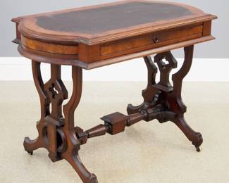 A 19th century American Victorian Period writing table. Walnut construction featuring a shaped top with inset faux leather surface and a single drawer at one side and a trestle base with ornately shaped supports and stretcher which rest on casters. Replaced faux leather top and wear to finish. 42 x 24 1/2 x 30 1/2" high overall. ESTIMATE $300-400