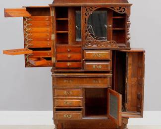 A turn of the century Fancy Oak Dental cabinet by "The Ransom Randolf Co., Toledo, Ohio, USA". Quarter sawn Oak construction with raised panel detail features a tall back with shaped upper shelf above a molded shelf supported by a turned spindle and fretwork facade in front of a sliding beveled glass door with a hinged side door with multiple swing out tool trays and three interior drawers, the lowest serving as a locking mechanism for the two short, one long and three short exterior doors at the front, includes another small door with beveled mirror and another swing out door on the right, open lower shelf and simple block feet. Original makers decal on left door interior. Original dark finish with wear and minor damage, lacks upper crest. 31 x 15 1/2 x 69" high overall. ESTIMATE $1,000-2,000