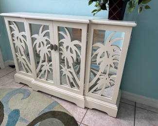 Beautiful coastal accent cabinet featuring decorative palm tree mirrored doors, an elegant white finish, and interior storage. Perfect for a foyer, dining room, living room, or Florida room with coastal, tropical, or beach-inspired décor.