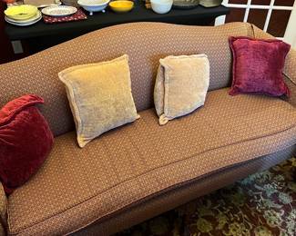 Camelback Sofa — This brown patterned sofa located in the front room offers a classic and comfortable seating option. The sofa features a gracefully curved back with nicely rolled arms, upholstered in a textured fabric with a geometric pattern. It includes four plush cushions in beige and deep red velvet providing both style and comfort. Measuring approximately 70 inches wide, 32 inches deep, and 34 inches high, this piece suits traditional or transitional decor. It is in good condition with some typical wear visible on the fabric consistent with regular use.