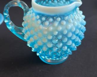 Blue Opalescent Hobnail Creamer — This charming blue opalescent hobnail creamer features a distinctive textured glass design that adds vintage farmhouse flair to any table setting. Crafted from pressed glass with a white opalescent top rim and a clear textured handle, it exudes timeless elegance and tactile appeal. Measuring approximately 4 inches tall by 5 inches wide including the handle, this creamer is the perfect size for vintage or antique dish collections. It is in excellent condition with no chips, cracks, or visible damage, preserving its classic beauty. Ideal as a collectible or functional accent piece for nostalgic glassware collectors.