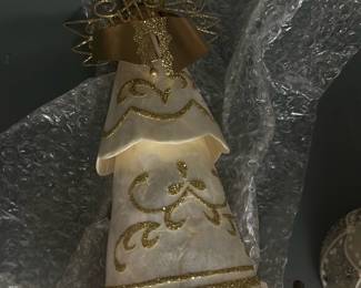 Gold Trim White shell angel — Decorative white angel figurine with gold glitter scrollwork and a metallic gold ribbon bow, ideal for holiday or year-round display. The cone-shaped layered dress appears to be a lightweight shell or resin with applied gold glitter accents and a wired halo/wing detail. Measures approximately 12 inches tall and is lightweight for easy placement; great for tabletop, mantel, or holiday vignette. In good condition with minor surface scuffs and a few small glue dots where decorative elements attach; currently wrapped in bubble wrap for storage/transport.