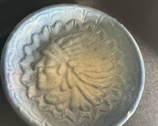 Vintage Native American — This vintage ashtray features a raised profile of a Native American chief in a full feather headdress. The ashtray has a light blue glaze with distinctive scalloped edges surrounding the central image. Measuring approximately 5 inches in diameter, it is a decorative yet practical collectible piece. The ashtray is in good vintage condition with some minor surface wear consistent with age. A unique accent for collectors of Native American or vintage Americana items.