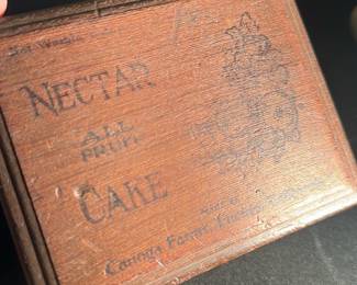 Nectar Cake Wooden Advertising Box — This vintage wooden advertising box features the inscription "Nectar All Fruit Cake" and branding from Carnegie Flavor Process, California. The small rectangular box is crafted from stained wood with dovetail joints and a hinged lid, perfect for display or storage of small items. The box measures approximately 3.5 inches in width, 2.5 inches in depth, and 2 inches in height, making it a compact collectible piece. It is in good condition with minor wear consistent with age, including some fading on the printed text and slight surface marks. A charming early 20th-century advertising collectible, ideal for nostalgia collectors or display in a vintage-themed setting.
