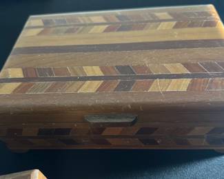 Wood Inlay Jewelry Box — This wood inlay jewelry box features a beautiful geometric pattern crafted from various wood tones, creating a distinctive decorative effect. The box is rectangular with a hinged lid and a small wooden latch for secure closure. Measuring approximately 3 inches tall, 8 inches wide, and 5 inches deep, it is a practical size for storing small collectibles or keepsakes. The box shows some surface wear and scratches consistent with age but remains sturdy and functional. Its handcrafted design and natural wood materials make it a charming vintage decorative piece suitable for a bonus up room or dresser.
