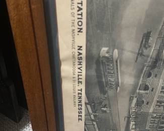 Union Station Nashville Print — Framed lithographic print depicting Union Station, Nashville, Tennessee (terminals of the Nashville, Chattanooga & St. Louis Railway). The piece is matted and set in a simple oak-tone wooden frame with glass, showing architectural detail of the historic station in grayscale. Overall size is moderate and suitable for wall display in a bonus room or office; frame shows light surface wear and the paper has mild age-toning and a small crease near the top edge. Good estate-sale condition for display with no major tears to the image; glass and frame are intact though the frame has minor scuffs consistent with normal use.