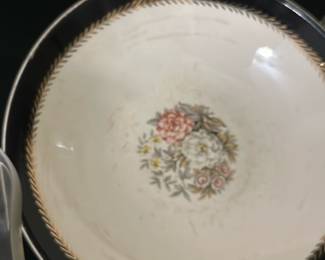 Vintage Floral Porcelain Dinner Plate — This vintage porcelain dinner plate features a delicate floral bouquet design in the center, accented by a gold laurel wreath border on a cream background. The plate has a black rim with gold trim that adds an elegant touch to its classic style. Measuring approximately 10.5 inches in diameter, it is suitable for both display and daily use. The plate shows some wear consistent with age, including minor surface scratches but no chips or cracks. This piece offers a charming addition to any vintage or traditional table setting.