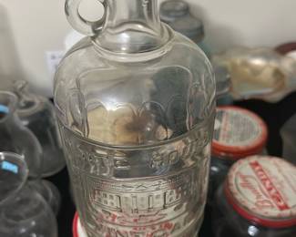 White House Vinegar Glass Bottle — This vintage White House Vinegar glass bottle features embossed lettering and a detailed depiction of the White House. Made of clear glass with a loop handle and cork stopper, it measures approximately 10 inches in height. The bottle is a charming collectible piece ideal for display or decorative use. It is in good condition, with minor wear consistent with age and no visible cracks or chips. This collectible reflects historical Americana branding and makes an excellent addition to vintage glassware collections.