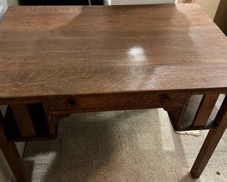 Oak Mission Style Writing Desk — 42” x 28”. This oak Mission style writing desk features a sturdy, rectangular top with characteristic straight legs and an open partitioned shelf space on either side. The rich oak finish highlights the wood grain, with a single center drawer for storage. Measuring approximately 30 in height, 46 in width, and 26 in length, it fits well in a bonus up room or office space. The desk is in good condition with minor surface wear typical of use over time. This practical piece reflects classic early 20th-century design aesthetics known for simplicity and craftsmanship.
