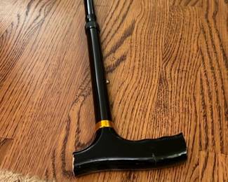 Black Adjustable Walking Cane — This black adjustable walking cane features a sturdy T-shaped handle with a gold accent ring for added style. Designed to provide support and stability, the cane is made from lightweight metal with an adjustable height mechanism. It measures approximately 36 inches in length and is suitable for everyday use. The cane is in good condition with light surface wear visible on the handle. Ideal for individuals seeking mobility assistance with a classic and practical design.