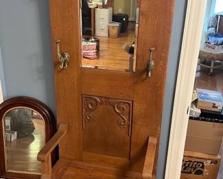 Carved Oak Hall Tree with Mirror — This carved oak hall tree features a tall back with a center mirror, four decorative brass hooks, and intricate floral carving accents. The warm oak finish highlights the detailed craftsmanship, complemented by a spacious bench seat and a lower storage drawer. Measuring approximately 72 inches high, 26 inches wide, and 16 inches deep, it fits ideally in a living room or entryway for functional storage and seating. The piece is in good condition with minor surface wear consistent with age, showing its vintage character. A practical and decorative vintage hall tree elevating any home's entrance or living area.