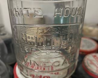 White House Vinegar Glass Bottle — This vintage White House Vinegar glass bottle features embossed lettering and a detailed depiction of the White House. Made of clear glass with a loop handle and cork stopper, it measures approximately 10 inches in height. The bottle is a charming collectible piece ideal for display or decorative use. It is in good condition, with minor wear consistent with age and no visible cracks or chips. This collectible reflects historical Americana branding and makes an excellent addition to vintage glassware collections.