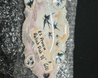 RS Prussia Pink Relish Dish 8-inch — This charming RS Prussia pink relish dish features delicate hand-painted blue swallows against a soft pink and cream background. The porcelain dish measures 8 inches in length and showcases ornate scalloped edges with a pierced handle for easy carrying. It is in good condition with minor signs of age and wear consistent with vintage porcelain. A lovely collector's piece from the RS Prussia line, ideal for display or special serving occasions.