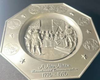 A New Nation Bicentennial Plate — This commemorative plate titled 'A New Nation Bicentennial Commemorative 1776 - 1976' honors the United States Bicentennial. It features a detailed embossed design of a historic scene with figures gathered around a table, framed by an octagonal border adorned with stars and circular historical vignettes. Crafted in metal with a dull silver finish, this piece is a collectible memorabilia item celebrating American history. Measuring approximately 8 inches in diameter, it is in good condition with minimal surface wear consistent with age. A meaningful collector’s piece from the 1976 U.S. Bicentennial celebration.