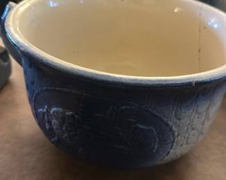 Blue Ceramic Stoneware Bowl — This blue ceramic stoneware bowl features a detailed embossed floral design around its exterior, adding vintage charm to any dining room setting. The bowl has a smooth cream-colored interior contrasting with the rich blue outer glaze. It measures approximately 5 inches in height, 8 inches in width, and 8 inches in length, making it a versatile piece for serving or display. There is visible wear and a fine crack around the rim, consistent with age, but no major damage that affects usability. Its sturdy construction and classic look make it a collectible vintage kitchen or dining accessory.