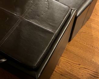 Black Faux Leather Storage Ottomans — These black faux leather storage ottomans offer a versatile combination of seating and hidden storage. Each ottoman features a hinged cushioned top with cross-stitched detailing and a spacious interior compartment for storing household items. Measuring approximately 16 inches in height, width, and length, these ottomans are lightweight and easy to move with built-in carry handles. The condition is good with some visible surface wear consistent with use but no major damage. Perfect for living room use as extra seating and convenient storage solution.