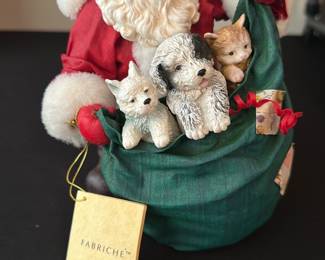 Fabriche KSA Santa with Pets — Charming Fabriche (KSA Collectibles) Santa figurine in a green sack filled with three small animal companions. Made of mixed materials with plush trim on Santa's red suit and detailed painted resin faces on the dog, cat and puppy; original Fabriche/KSA hang tag attached. Measures approximately 12 inches tall overall and is a tabletop holiday accent for mantel or entryway displays. Good condition with light shelf wear and intact fabric; tag present indicating collectible line.