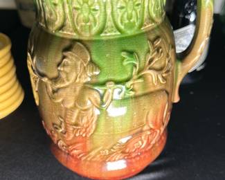 Green and Orange Ceramic Pitcher — This charming ceramic pitcher features a detailed relief design depicting a man playing a horn. The pitcher is glazed in vibrant green at the top, blending into warm orange at the base, with an intricate floral pattern adorning the rim. It measures approximately 8 inches in height and 6 inches in width, ideal for decorative display or functional use. The piece is in good condition with minor wear consistent with age, and no visible cracks or chips. Its unique design and warm colors make it an attractive addition to any vintage or rustic collection.
