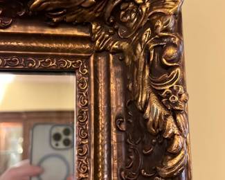 Ornate Gold Leaf Wall Mirror — This ornate gold leaf wall mirror features an intricately carved frame with detailed floral and scroll motifs. The large rectangular mirror is perfect for adding elegance and light to any dining room or living space. Measuring approximately 36 inches in height and 48 inches in width, it is a substantial decorative piece. The mirror and frame are in good condition with minor wear consistent with its vintage style. Its classic design makes it a timeless accent for traditional or vintage-inspired interiors.